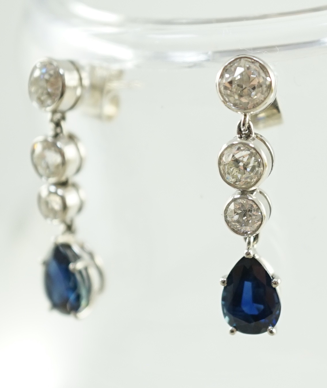 A pair of 1980's platinum, single stone pear cut sapphire and graduated three stone collet set diamond drop earrings